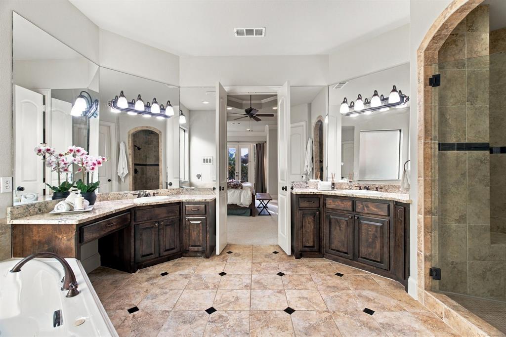 Dual vanities with stone countertops, stone-tile flooring with decorative inserts, built-in soaking tub, and a walk-in shower with arched tile surround