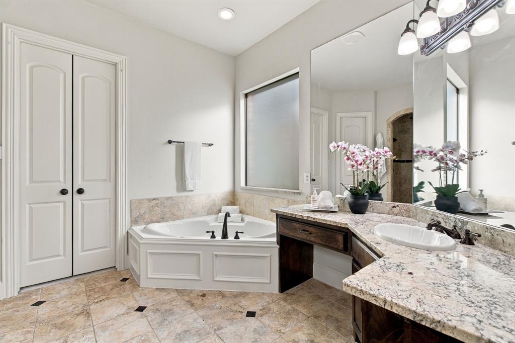 Corner soaking tub with tile surround, stone-finish vanity countertop with an integrated oval basin, full-width vanity mirror, dark wood-finish cabinetry, and ceramic tile flooring with decorative inlay accents