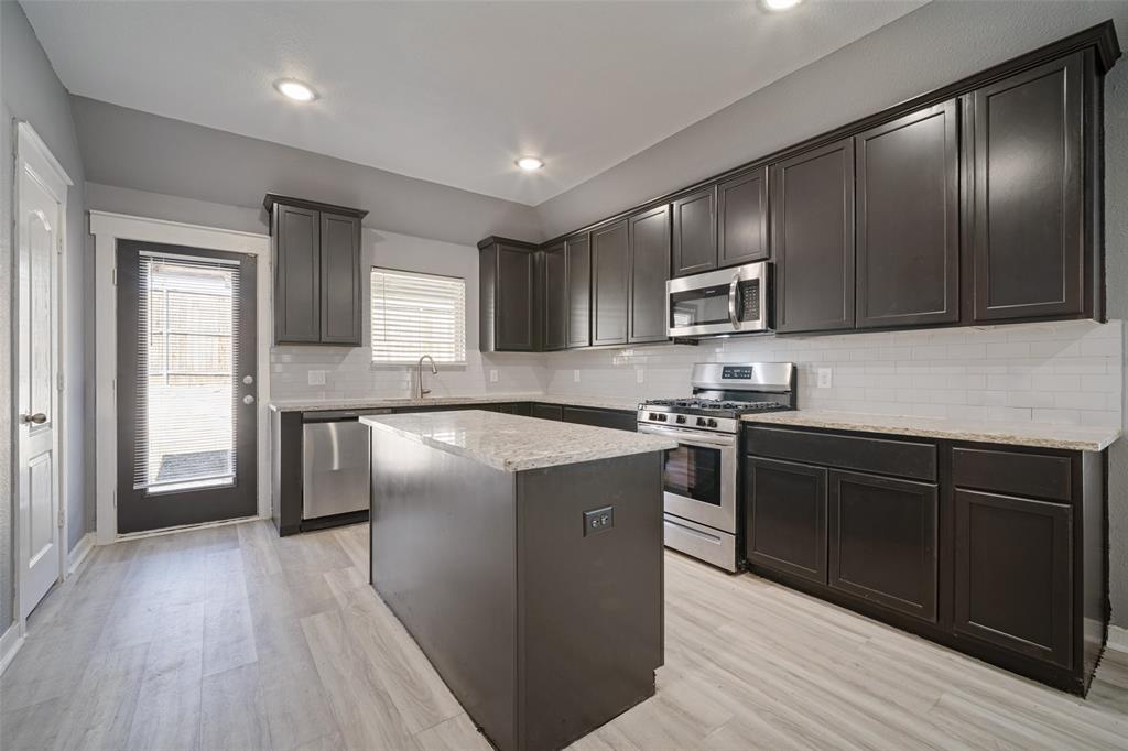 Kitchen featuring dark-finish cabinetry, light-toned stone-style countertops, white subway tile backsplash, stainless steel appliances, and wood-finish flooring Kitchen featuring dark-finish cabinetry, light-toned stone-style countertops, white subway tile backsplash, stainless steel appliances, and wood-finish flooring