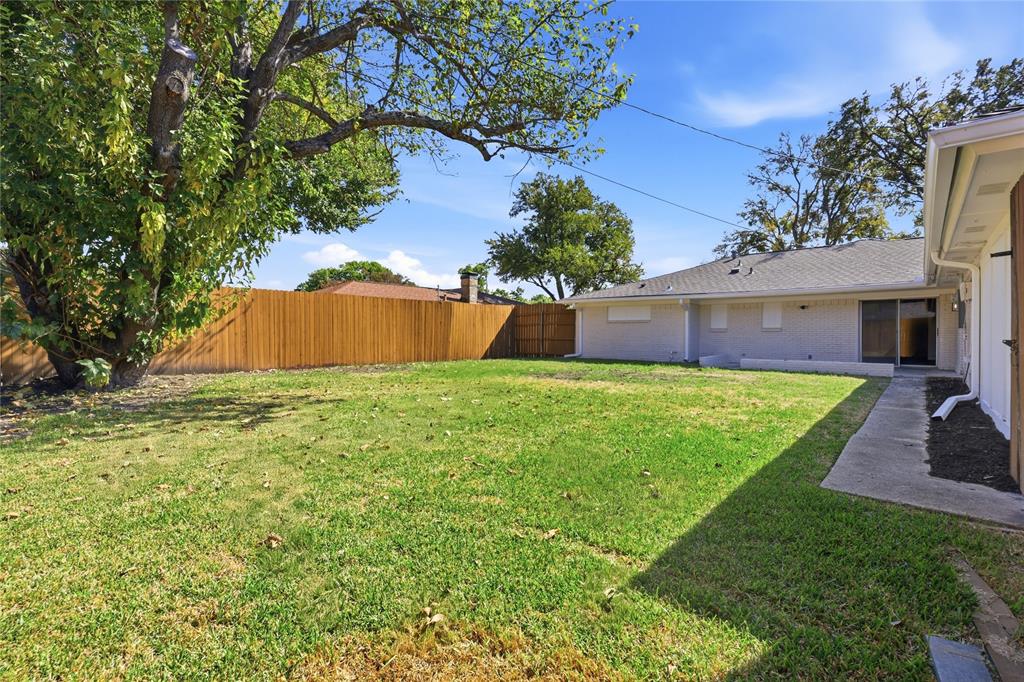 Backyard featuring a grass lawn, wood privacy fencing, mature trees, concrete pathway, and light-colored brick exterior