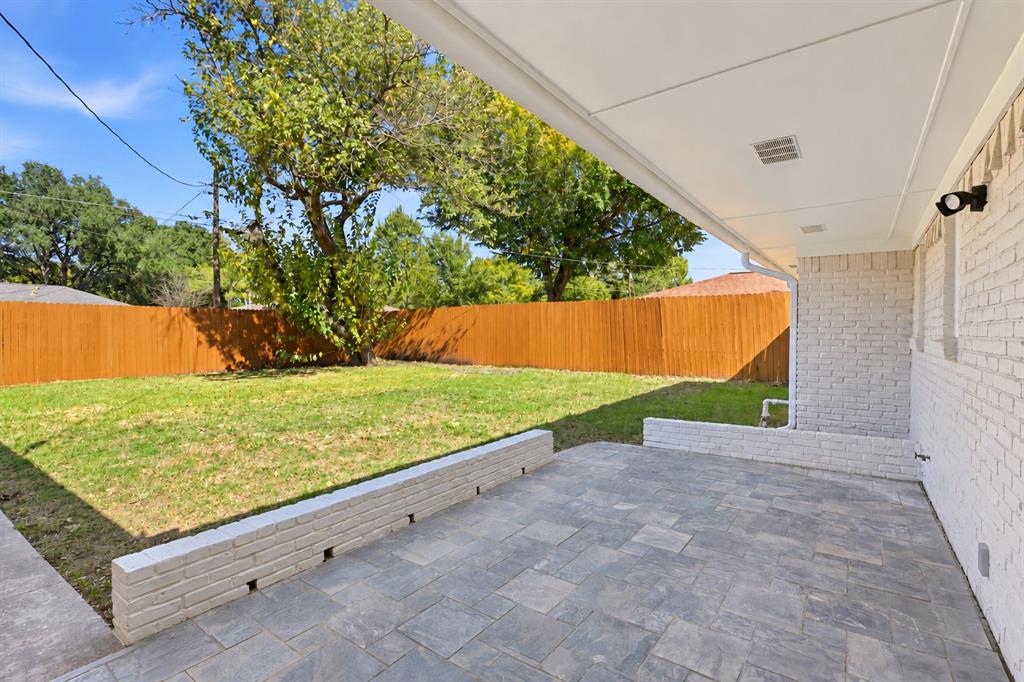 Covered patio featuring stone-tiled flooring, a built-in brick retaining wall, a manicured lawn, and perimeter wood privacy fencing