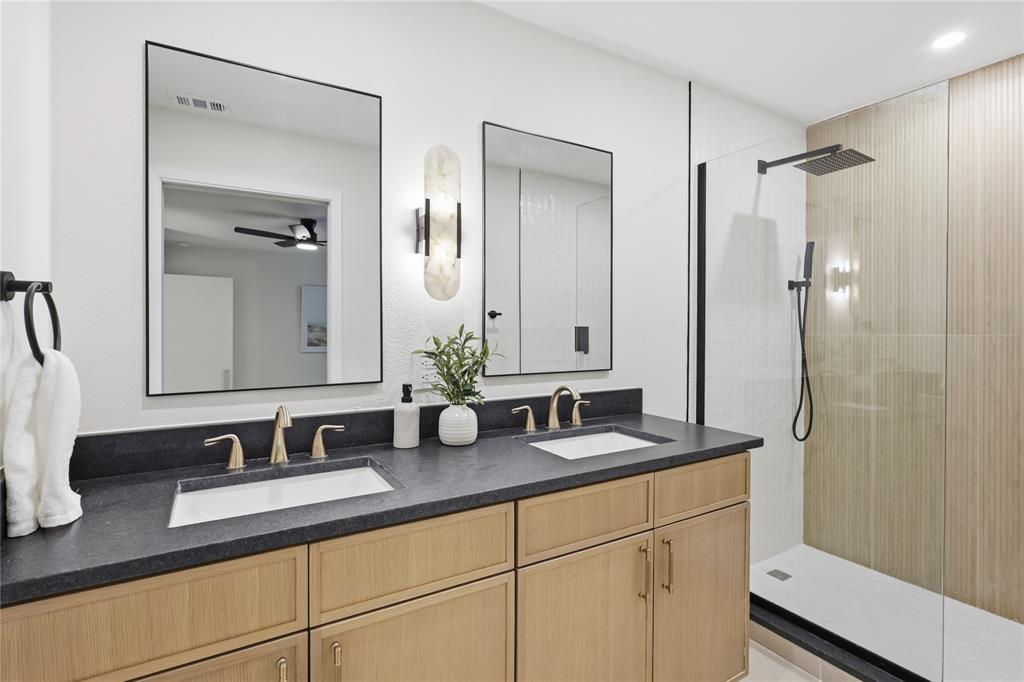 Double vanity featuring wood-finish cabinetry, dark solid-surface countertops, rectangular undermount sinks, gold-tone faucets, and two frameless mirrors with a vertical wall-mounted light fixture