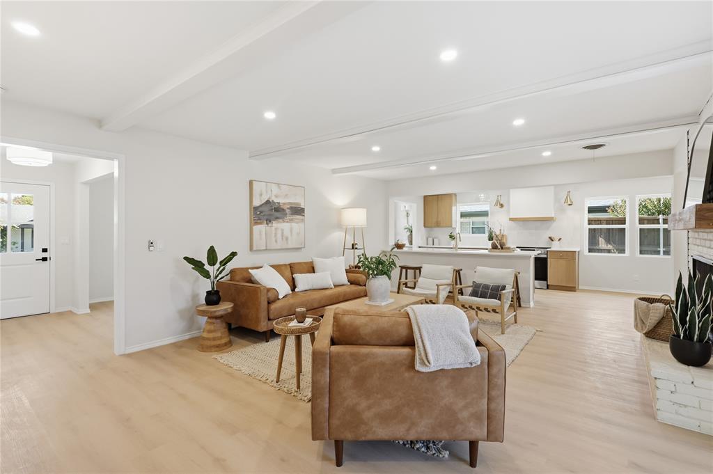 Open-concept interior featuring light wood-finish flooring, recessed lighting, exposed ceiling beams, a kitchen breakfast bar, and a painted brick fireplace surround Open-concept interior featuring light wood-finish flooring, recessed lighting, exposed ceiling beams, a kitchen breakfast bar, and a painted brick fireplace surround