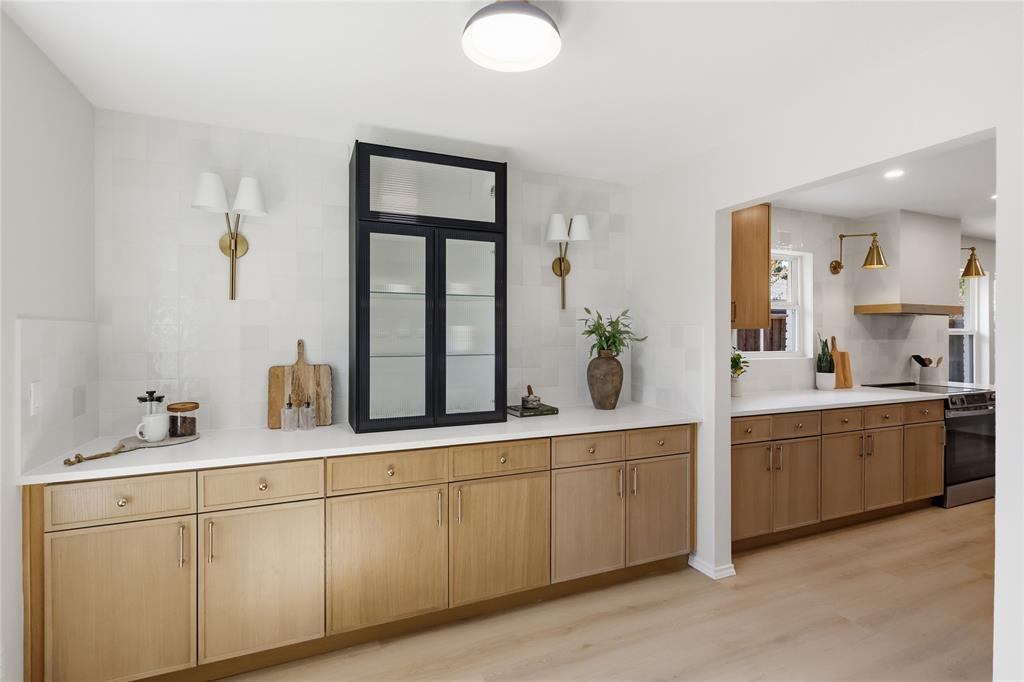 Kitchen featuring custom light-wood cabinetry, white solid-surface countertops, integrated glass-front display cabinetry, brass-toned wall sconces, and wood-finish flooring