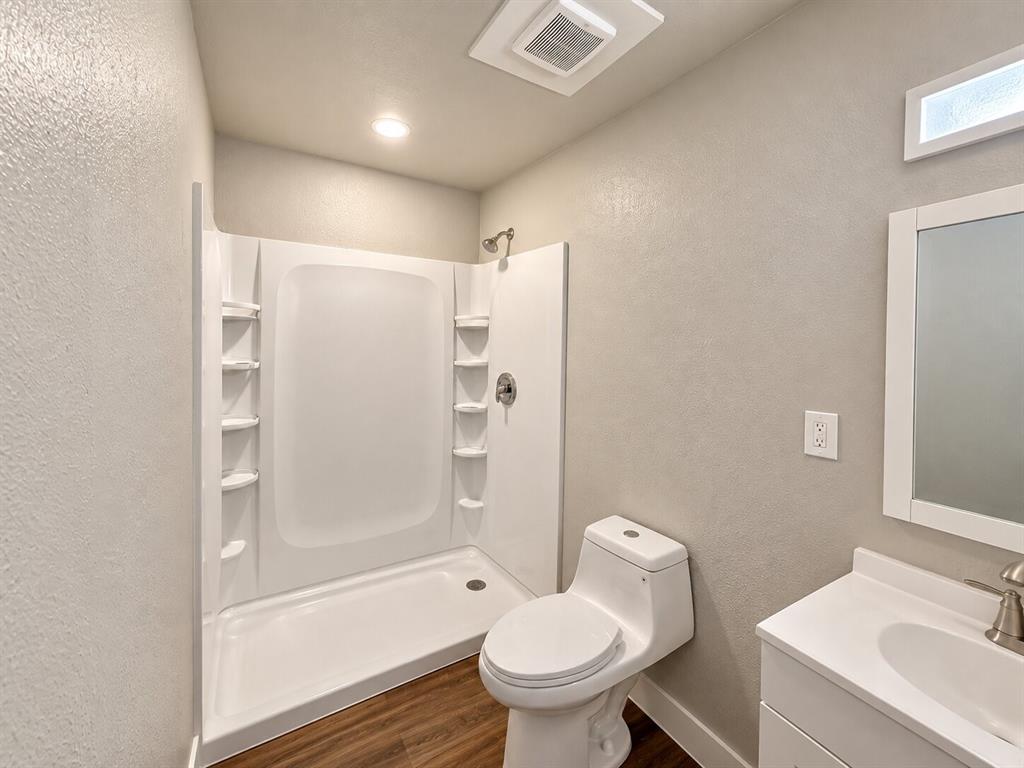 White shower stall with built-in shelving, wood-finish flooring, white toilet, single vanity with white countertop, and brushed nickel fixtures White shower stall with built-in shelving, wood-finish flooring, white toilet, single vanity with white countertop, and brushed nickel fixtures