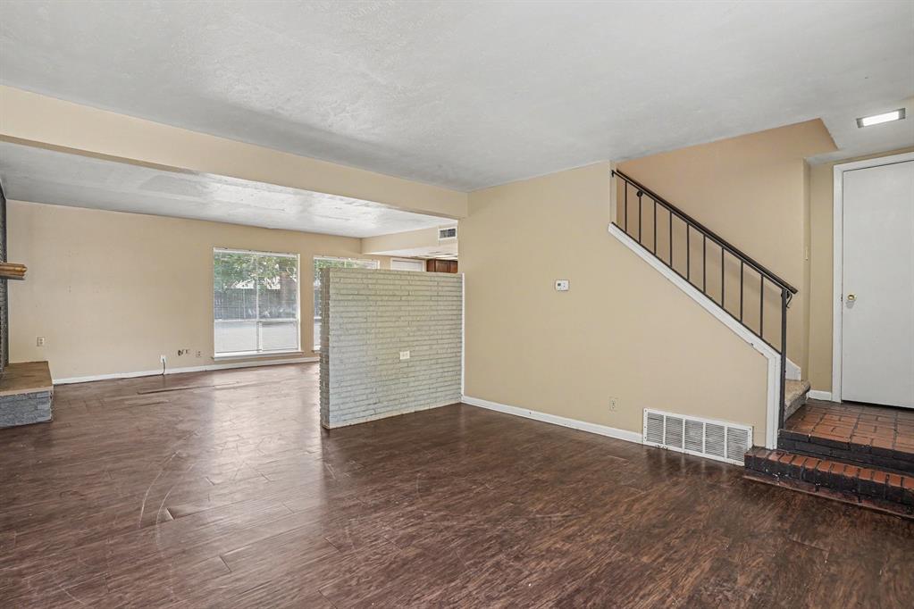 Wood-finish flooring throughout, brick accent partition, fireplace with stone surround, large window with outdoor visibility, and a staircase with wrought iron railing