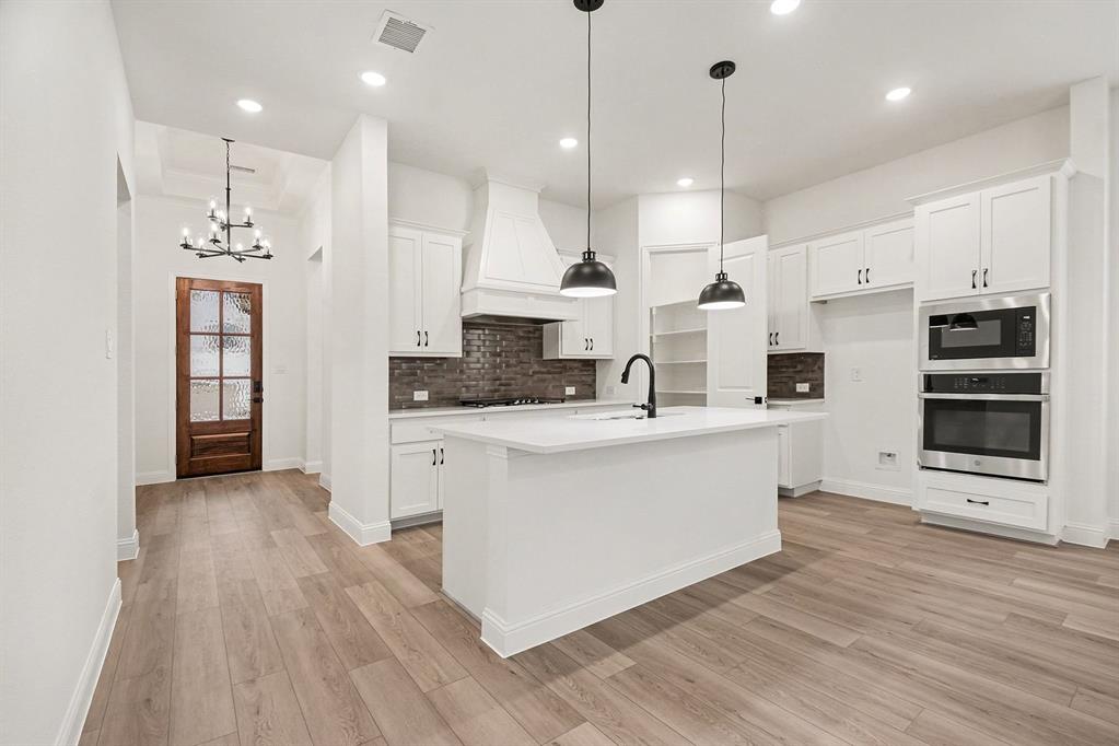 Kitchen featuring white shaker-style cabinetry, central island with undermount sink, brick-style backsplash, integrated double wall ovens, and wood-finish flooring Kitchen featuring white shaker-style cabinetry, central island with undermount sink, brick-style backsplash, integrated double wall ovens, and wood-finish flooring