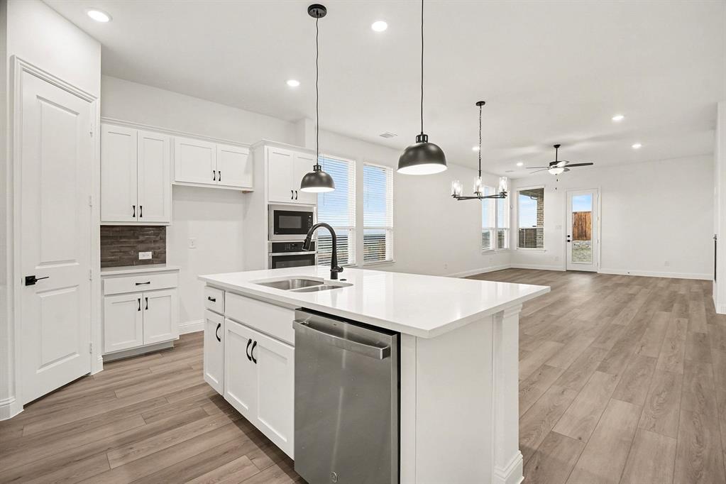 Kitchen featuring a central island with a solid surface countertop, white shaker-style cabinetry, stainless steel built-in appliances, pendant lighting, and wood-finish flooring Kitchen featuring a central island with a solid surface countertop, white shaker-style cabinetry, stainless steel built-in appliances, pendant lighting, and wood-finish flooring