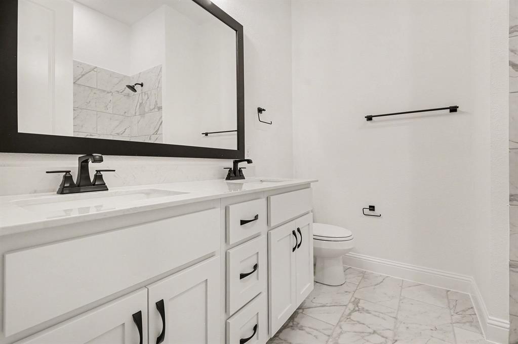 Double vanity with white cabinetry, stone-finish countertops, matte black fixtures, framed mirror, and stone-tile flooring