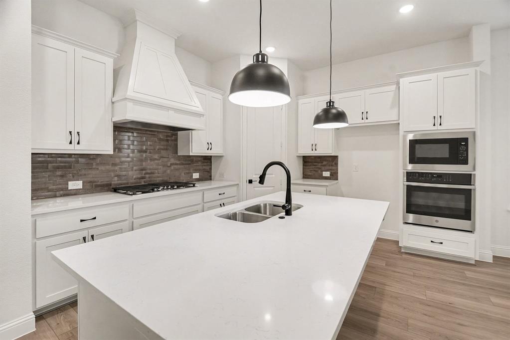 Kitchen featuring a large central island with light-toned solid surface countertops, custom white cabinetry, built-in stainless steel double ovens, tile backsplash, and matte black pendant lighting Kitchen featuring a large central island with light-toned solid surface countertops, custom white cabinetry, built-in stainless steel double ovens, tile backsplash, and matte black pendant lighting
