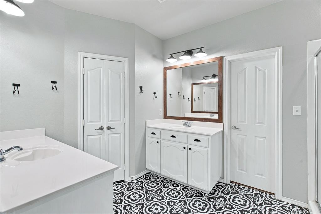 Bathroom featuring dual white vanities with solid-surface countertops, pattern-tiled flooring, three-light wall-mounted fixtures, and wood-framed mirrors