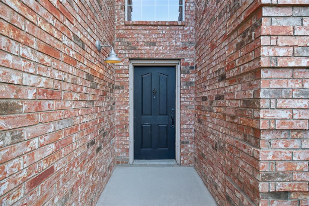 Exterior entryway featuring red brick facade, four-panel door, metal wall-mounted light fixture, and concrete porch
