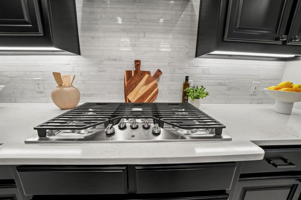 Stainless steel gas cooktop with heavy-duty grates, light-toned stone-finish countertops, linear tile backsplash, and dark shaker-style cabinetry with under-cabinet lighting