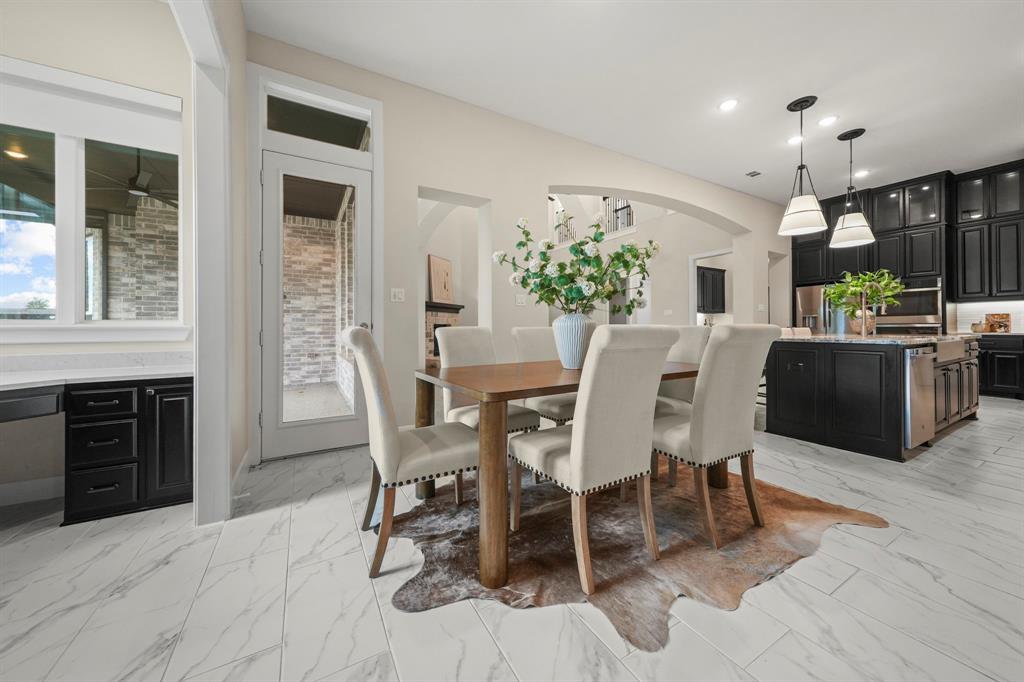Open-concept kitchen and dining area featuring stone-look tile flooring, shaker-style cabinetry, a central island with an integrated sink, dual pendant lighting, and arched architectural details