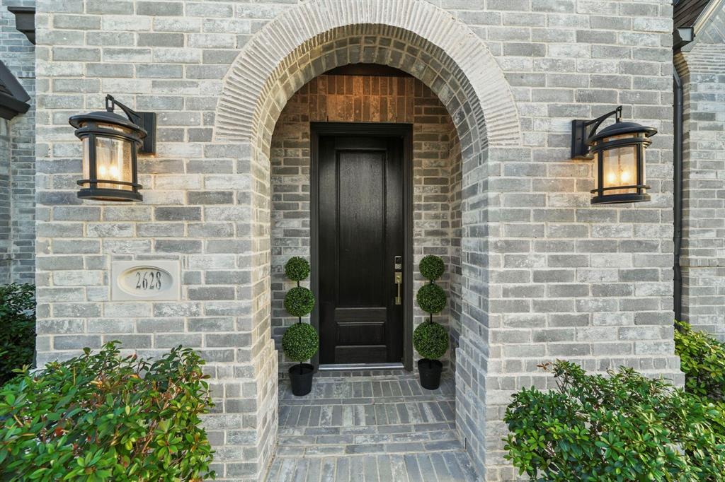 Entryway featuring a grey brick facade, arched stone surround, solid dark-finish entry door, and two wall-mounted metal lanterns
