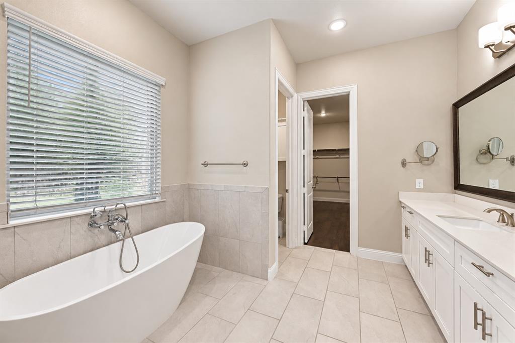 Freestanding soaking tub with wall-mounted faucet, dual vanity with undermount sinks, stone countertop, and tile flooring