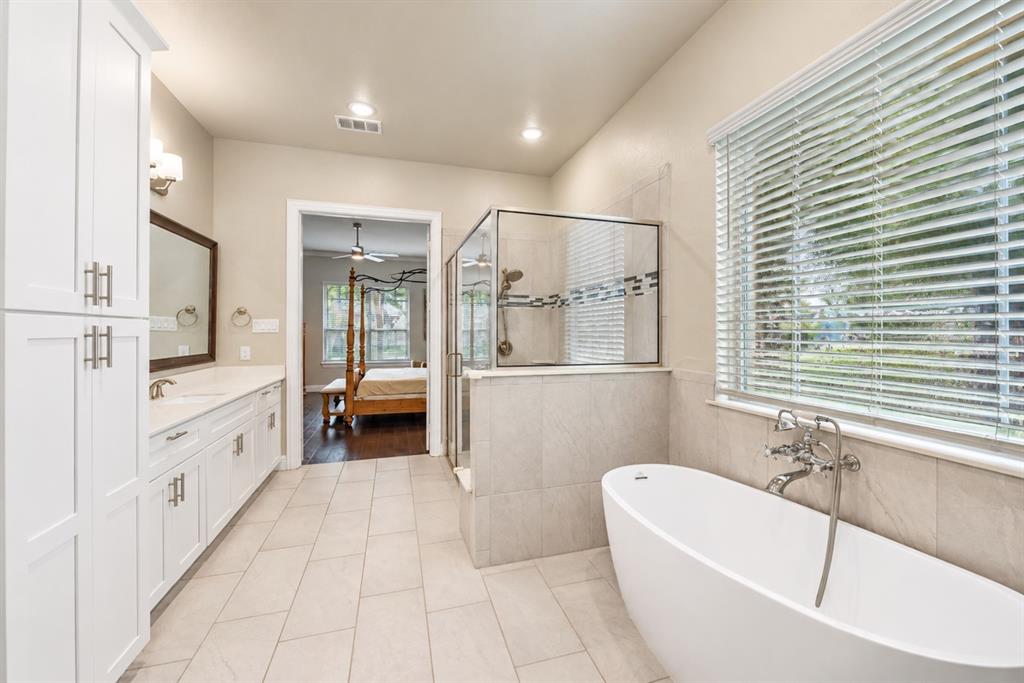 Freestanding soaking tub, walk-in shower with glass enclosure, dual vanity with white cabinetry, and large window with blinds