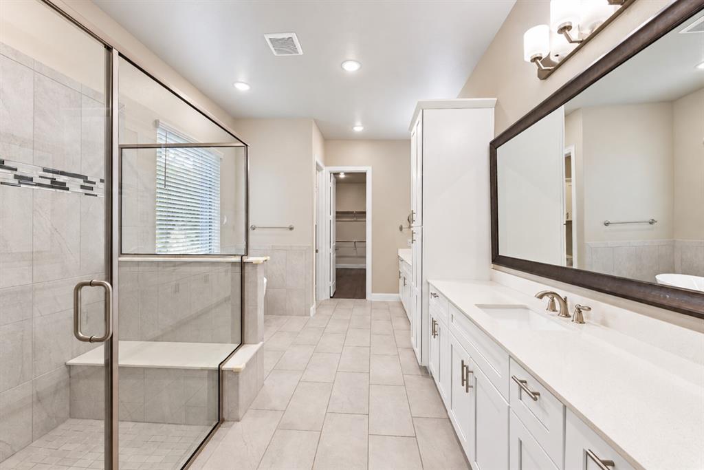 Glass-enclosed shower with integrated bench seating, dual vanity with undermount sinks, and a full-width mirror with decorative lighting
