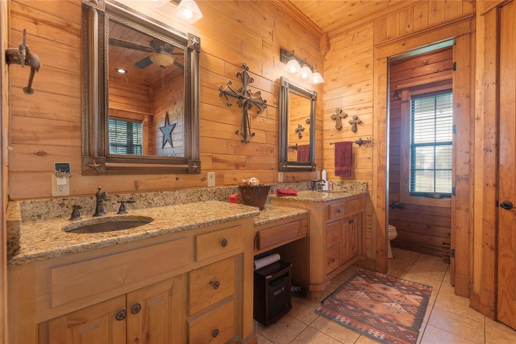 Dual-sink vanity with natural stone countertops, under-mount sinks, wood cabinetry, and framed mirrors with integrated lighting, finished with tile flooring and wood-paneled walls