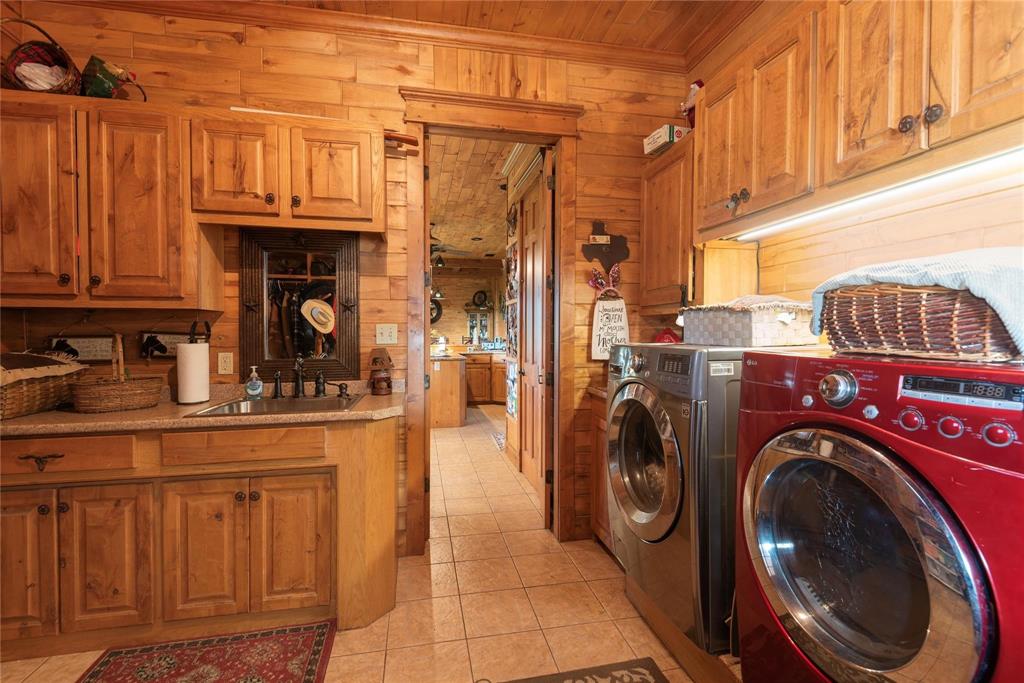 Laundry room featuring knotty wood wall paneling, custom wood cabinetry, stone-finish countertops, an under-mount stainless steel sink with a bronze-finish faucet, and ceramic tile flooring