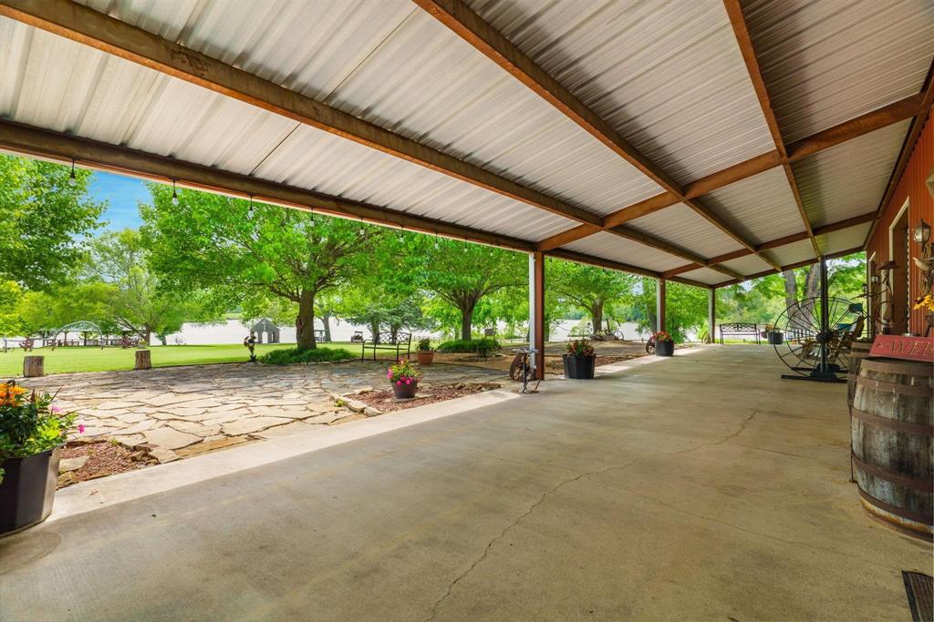 Covered concrete patio featuring an exposed-beam ceiling with corrugated metal roofing, stone hardscaping, and mature trees