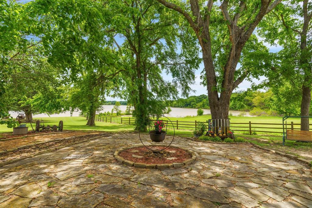 Flagstone patio featuring a circular stone inlay, mature hardwood trees, grass lawn, wood-post fencing, and water frontage