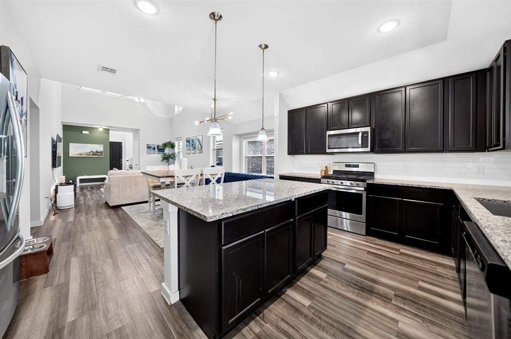 Open-concept kitchen featuring dark wood-finish cabinetry, light-toned granite countertops, a subway tile backsplash, stainless steel appliances, and wood-finish flooring
