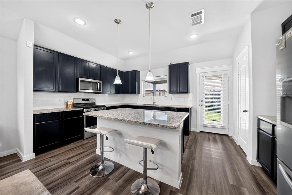 Modern kitchen featuring black cabinetry with stainless steel appliances, white quartz countertops, a large waterfall-edge island with seating for two, pendant lighting, recessed ceiling lights, and wide-plank wood-finish flooring throughout