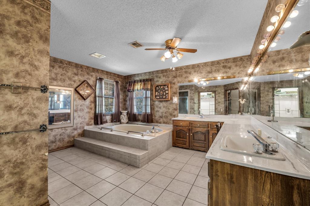 Tile flooring, built-in soaking tub with tiled surround, dual vanity with undermount sinks, wood-finish cabinetry, and mirrored wall with integrated lighting
