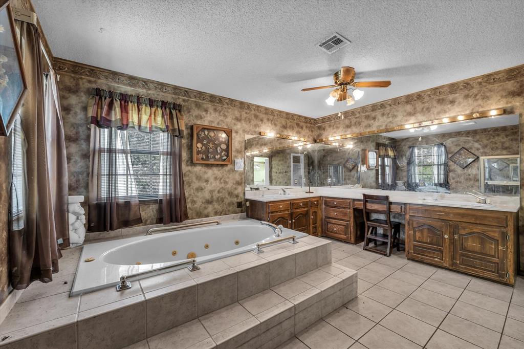 Built-in jetted tub with tiled surround, dual vanity with wood cabinetry, large vanity mirror with integrated lighting, and ceiling fan