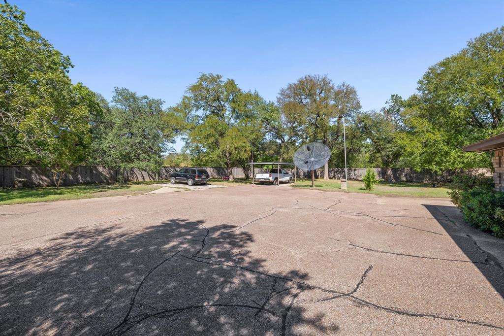 Paved surface, mature trees, partial fencing, and a covered parking structure