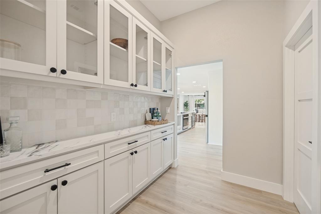 White shaker cabinetry with black hardware, glass-front upper cabinets, white tile backsplash, light wood-finish flooring, and a white countertop with gray veining