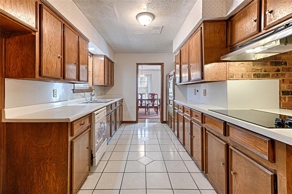 Galley kitchen featuring wood cabinetry, white solid-surface countertops, tile flooring, built-in wall oven, and a brick-accented cooktop surround Galley kitchen featuring wood cabinetry, white solid-surface countertops, tile flooring, built-in wall oven, and a brick-accented cooktop surround