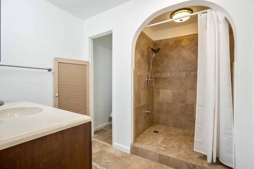 Bathroom featuring a walk-in shower with stone-finish tile surround and flooring, an arched alcove, a vanity with a solid-surface countertop, and a built-in louvered-door cabinet