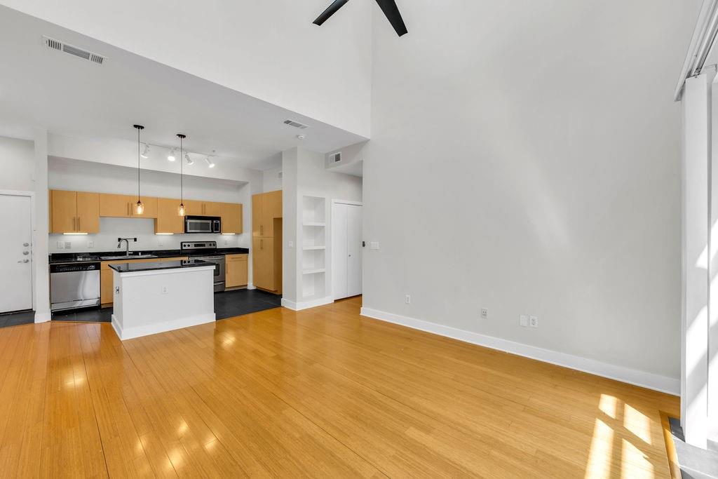 Open-concept living area with high ceilings, wood-finish flooring, and a kitchen featuring stainless steel appliances, dark countertops, and light wood cabinetry