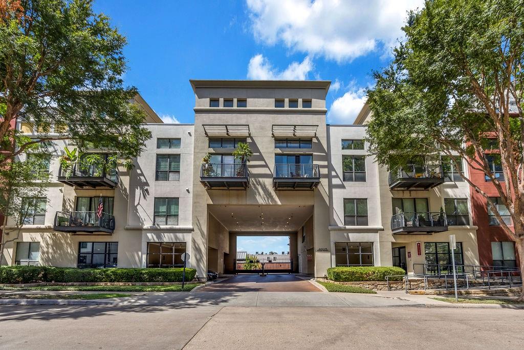 Multi-story building featuring a stucco exterior, multiple private balconies with metal railings, a central drive-through passage, and integrated landscaping