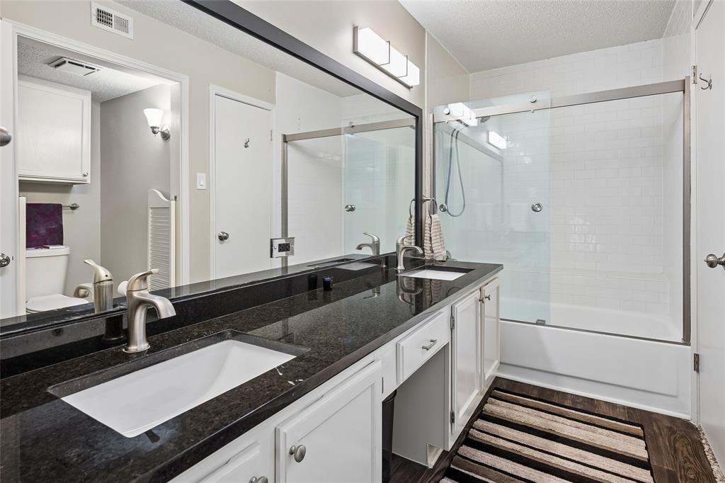 Dual-sink vanity with dark stone countertop, white cabinetry, full-width mirror, and shower-tub combination featuring a sliding glass door and tile surround