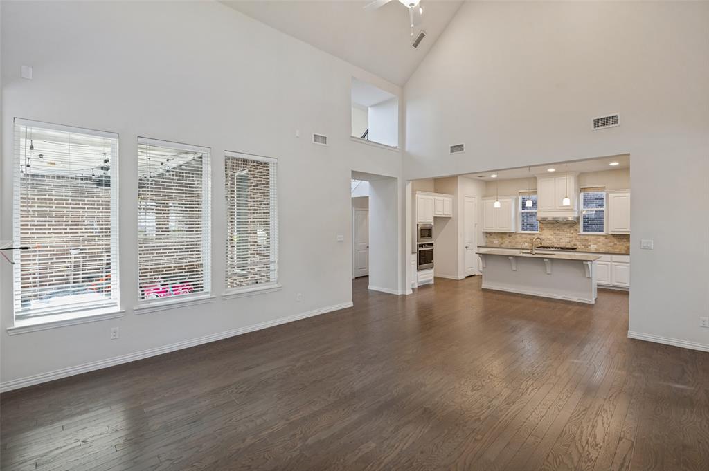 Expansive living area with vaulted ceilings, wood-finish flooring, three oversized windows, and an island kitchen featuring white cabinetry and built-in appliances Expansive living area with vaulted ceilings, wood-finish flooring, three oversized windows, and an island kitchen featuring white cabinetry and built-in appliances