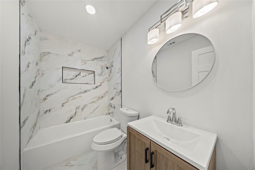 Bathroom featuring stone-finish wall tile, integrated shower niche, wood-finish vanity cabinet, oval wall mirror, and three-light vanity fixture