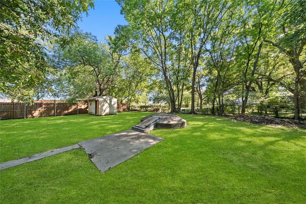 Expansive backyard featuring mature trees, a wood privacy fence, a detached storage structure, and a concrete storm shelter