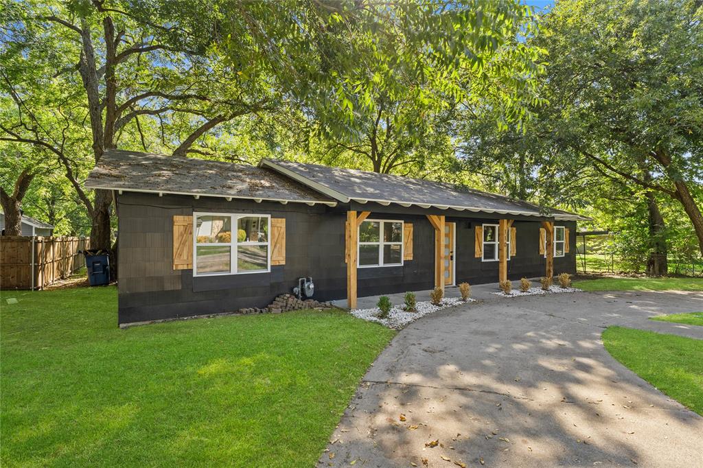 Single-story exterior featuring dark siding, wood-finish shutters, a covered front porch with timber support beams, and a concrete driveway surrounded by mature trees
