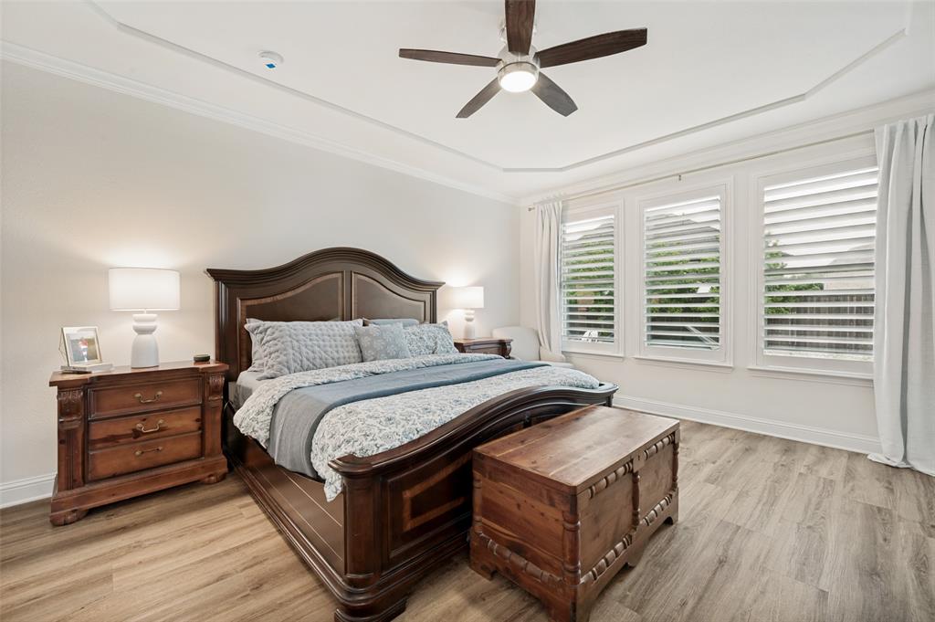 Tray ceiling with crown molding, ceiling fan, three windows with plantation shutters, wood-finish flooring, and white baseboards