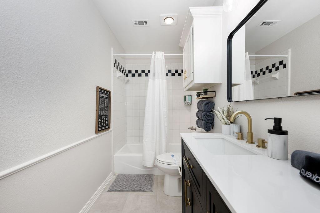 White tile shower with checkerboard accent, single vanity with white countertop, brass finish faucet, and recessed lighting