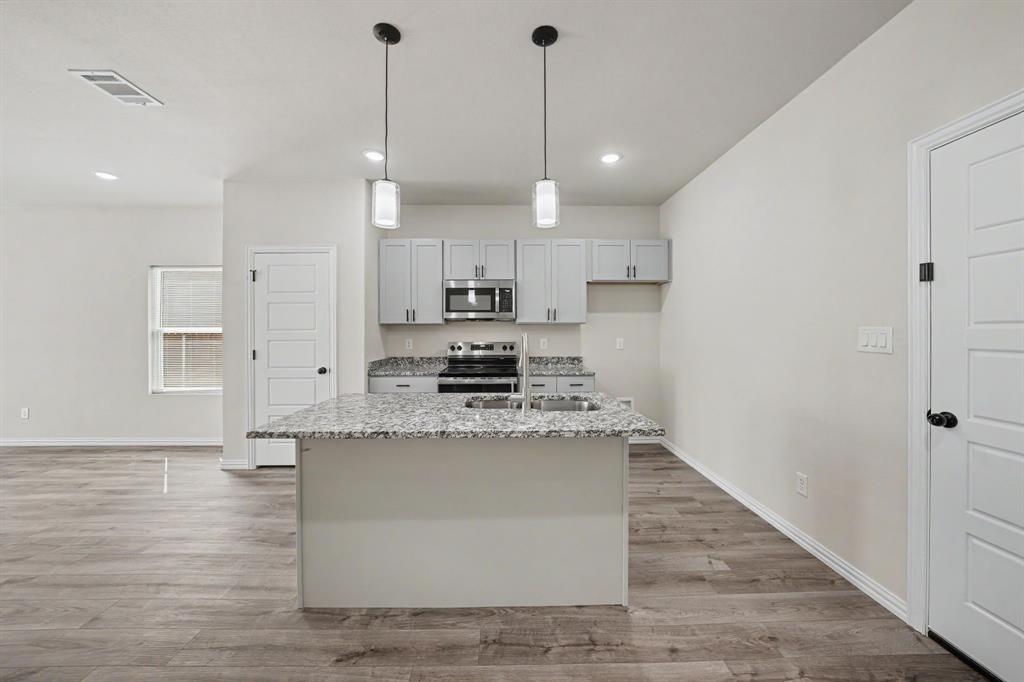 Kitchen featuring an island with a built-in sink, pendant lighting, light gray cabinetry, stainless steel appliances, and wood-finish flooring