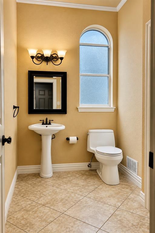 Pedestal sink, arched frosted window, tiled flooring, wall-mounted light fixture, crown molding