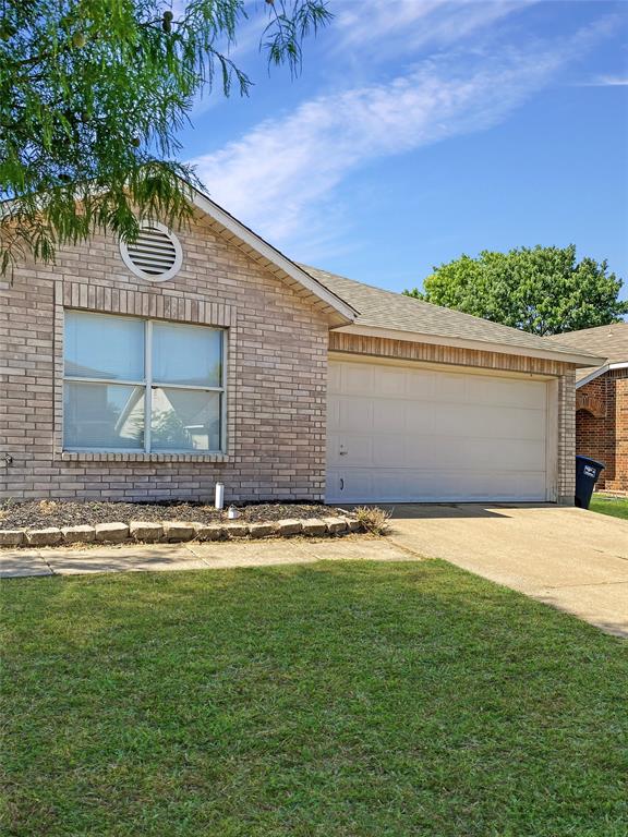 Charming brick home featuring a spacious two-car garage with paneled door, large front-facing window with contemporary framing, manicured lawn, and mature landscaping including trees and shrubs