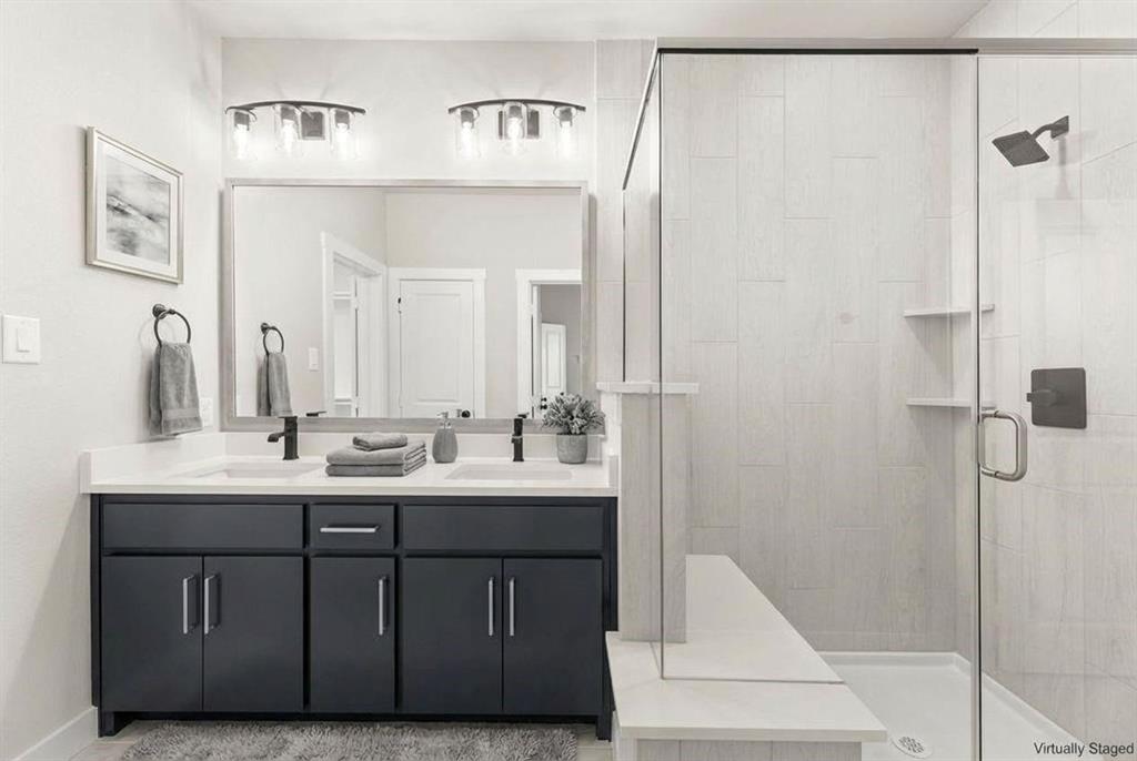 Dual-sink vanity with dark cabinetry and white countertop, full-width mirror with dual overhead lighting, and walk-in shower featuring glass enclosure and stone-finish wall tile