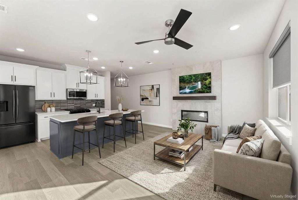 Open-concept living featuring a white cabinetry kitchen with black stainless steel appliances and a waterfall-edge island with seating, hardwood-finish flooring throughout, recessed and pendant lighting, ceiling fan, and an adjacent fireplace with built-in shelving and a wall-mounted television