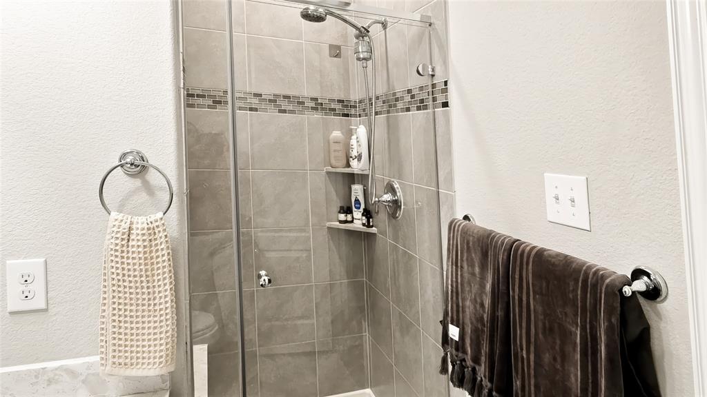 Walk-in shower featuring glass door enclosure, neutral-tone wall tiling, decorative horizontal tile inlay, integrated corner shelving, and a handheld shower fixture
