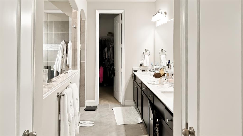 Dual-sink vanity with dark cabinetry, light stone-surface countertops, tile flooring, and walk-in closet with built-in shelving